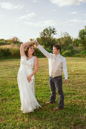 Dancing couple in wedding attire twirl and laugh together in a sunny green field.
