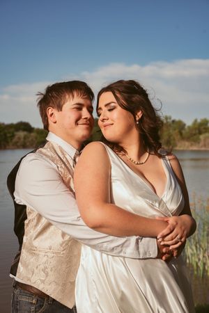A romantic wedding couple embraces by a lake during sunset in casual formal attire with vest and white dress.