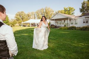 Wedding couple walks hand in hand across sunny green lawn with houses in background.