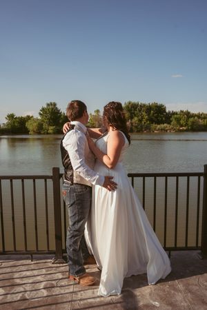 A couple embracing on a wooden deck overlooking a serene lake under blue skies in late afternoon light.