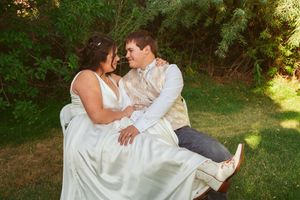 Two people in wedding attire share a romantic embrace in a sunlit green garden setting.