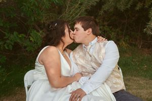 A romantic couple shares a tender kiss while sitting together outdoors in formal attire against a natural backdrop.