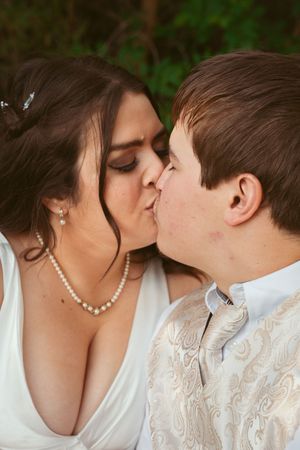 A tender kiss shared between newlyweds, with the bride wearing a pearl necklace and white dress against a green backdrop.