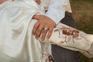 A hand wearing an engagement ring rests on a white dress near ornate embroidered floral designs.