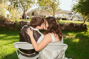 A couple shares a romantic kiss while sitting on white chairs in a sunny backyard garden setting.