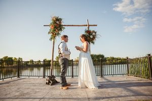 Outdoor lakeside wedding ceremony with decorative cross arch and couple standing on wooden deck at sunset.