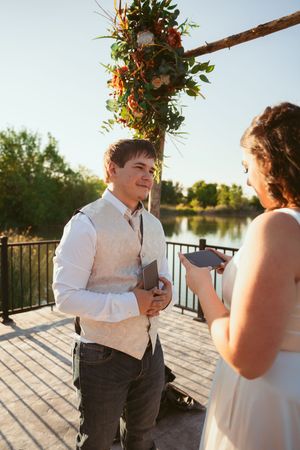Wedding ceremony on a lakeside deck with decorative floral arch during golden hour.