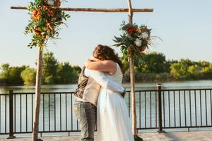 A couple embraces under a rustic wooden wedding arch with floral arrangements overlooking a serene lake at sunset.