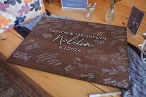 A wooden wedding guest book board with white signatures and messages surrounding the couple's names and date.