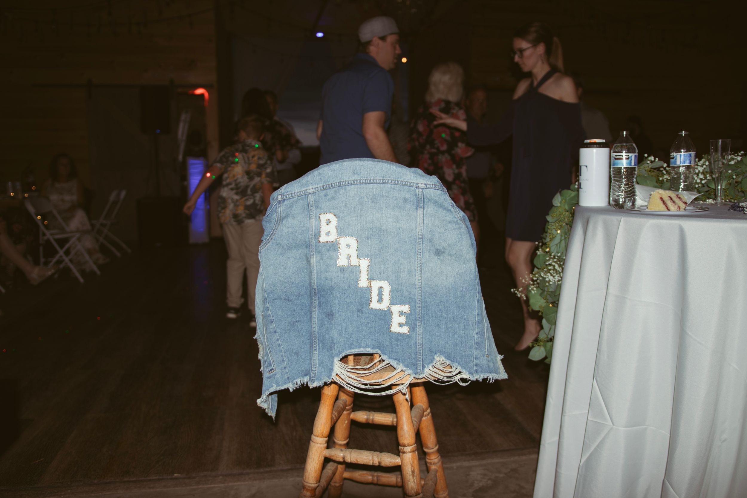 A decorated denim jacket with 'BRIDE' text hangs on a wooden stool at a dimly lit reception event.