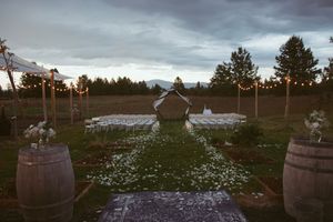 Rustic outdoor wedding ceremony setup with wooden barrels and white petals scattered along a grass aisle at dusk.