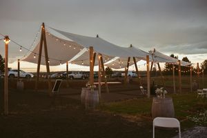 An outdoor event space with string lights and white canopy tents set up at dusk with empty chairs underneath.