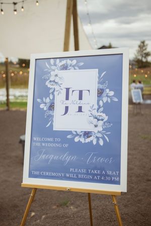 Blue and white floral wedding welcome sign on an easel with string lights in background.