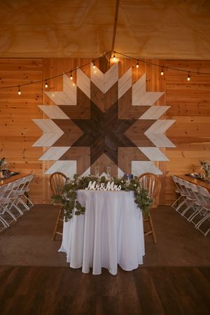 Rustic wedding reception setup with geometric wooden wall art and white sweetheart table decorated with greenery.