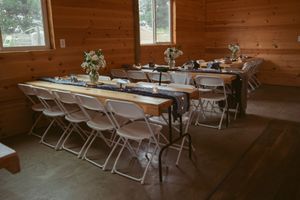 Long banquet tables with white linens and folding chairs set up inside a rustic wooden cabin reception venue.