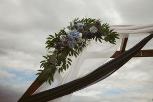 Floral wedding arch decoration with blue flowers and greenery against a cloudy sky.