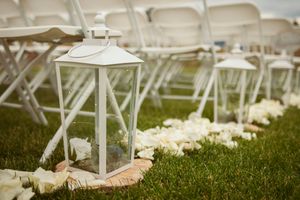 White decorative lantern and rose petals line an outdoor ceremony aisle with folding chairs.