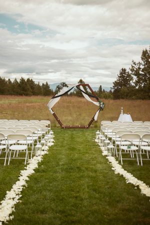 Outdoor wedding ceremony setup with wooden hexagonal arch and white chairs lined with flower petals.