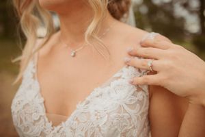 Close-up of lace wedding dress detail with delicate necklace and engagement ring.
