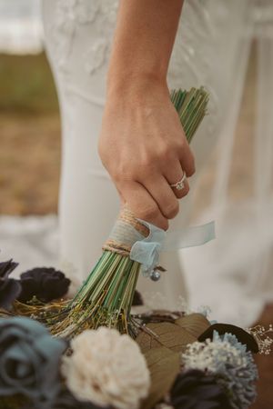 Hand holding a rustic bouquet wrapped in twine and blue ribbon showing engagement ring.