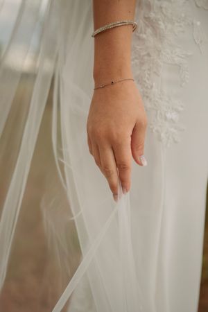 A bride's hand with delicate silver bracelets touches flowing white tulle fabric.