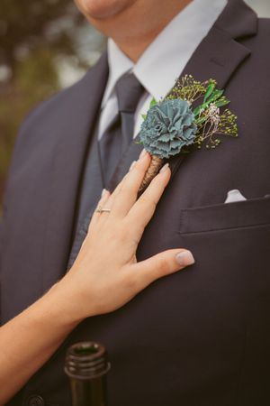 Close-up of hands adjusting a blue-green succulent boutonniere on a dark suit lapel.