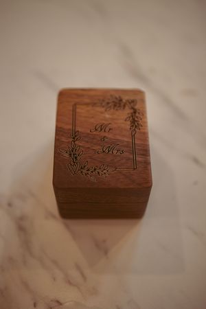 A small wooden ring box with floral engravings and 'Mr & Mrs' text sits on a marble surface.