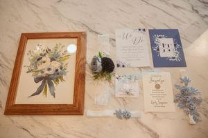 Wedding invitation suite displayed with framed floral artwork and blue flower accents on marble surface.