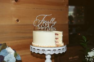 White wedding cake with 'Finally Mr & Mrs' topper displayed on an elegant lace cake stand against wooden backdrop.