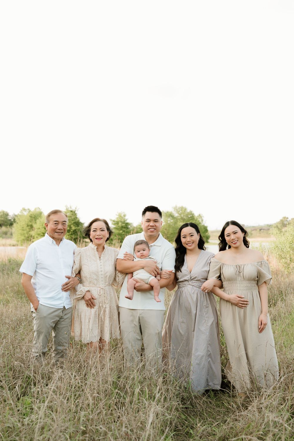 Extended family group in neutral colored outfits poses together in a grassy field at sunset.