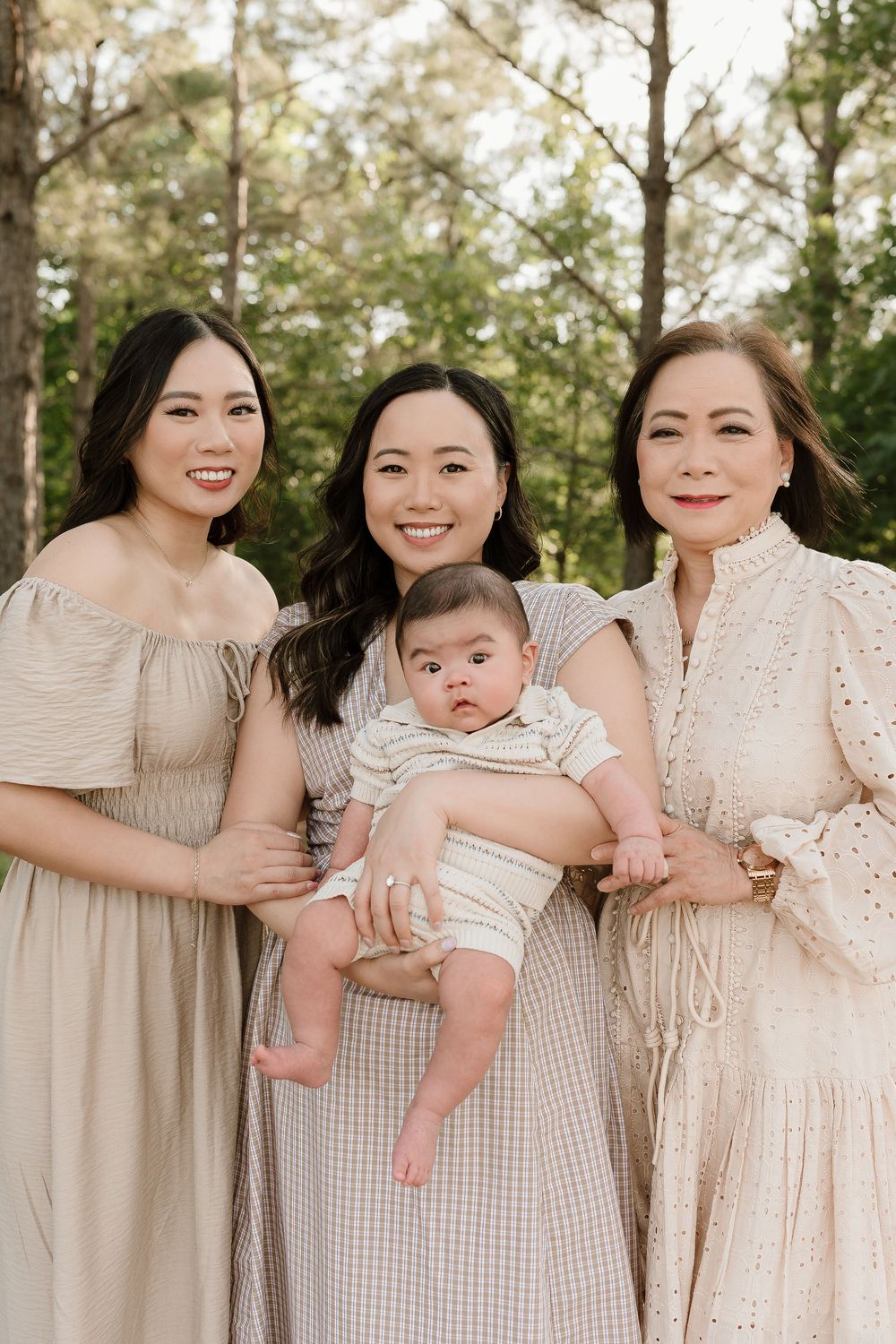 A group wearing coordinated cream-colored dresses poses together with a baby outdoors in a natural setting.