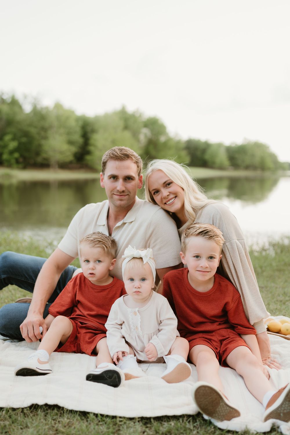 Family relaxing together on white blanket for outdoor portrait session by lake.