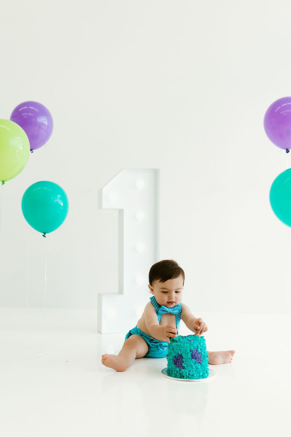 Baby in turquoise outfit with birthday cake and colorful balloons in white studio setting.