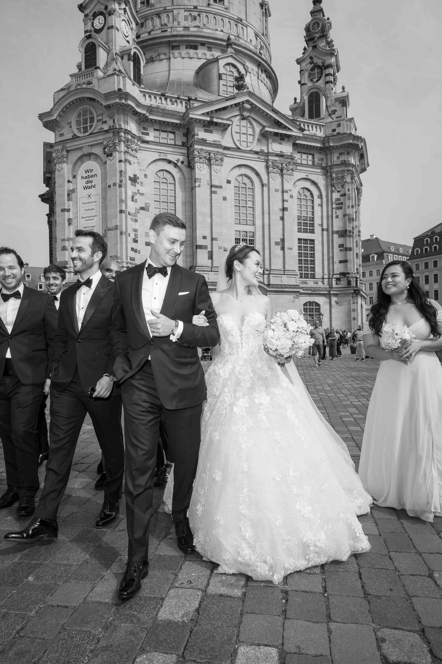 A wedding party walking together outside a grand baroque cathedral with ornate stone architecture in black and white.