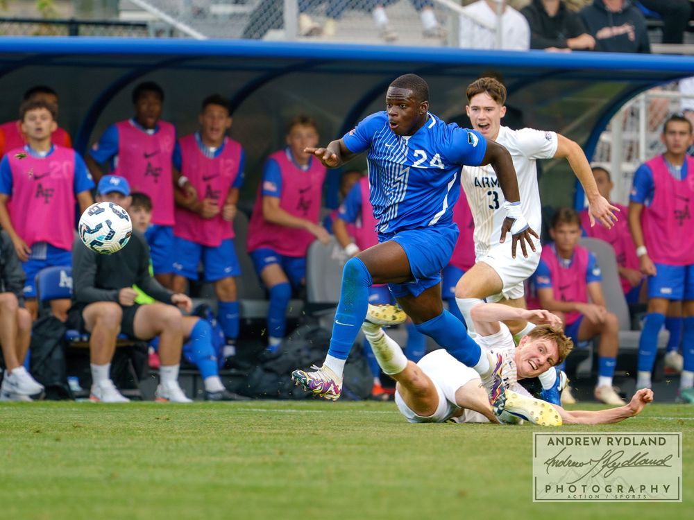 Soccer players battle for possession during a competitive match with pink-shirted substitutes watching from the bench.