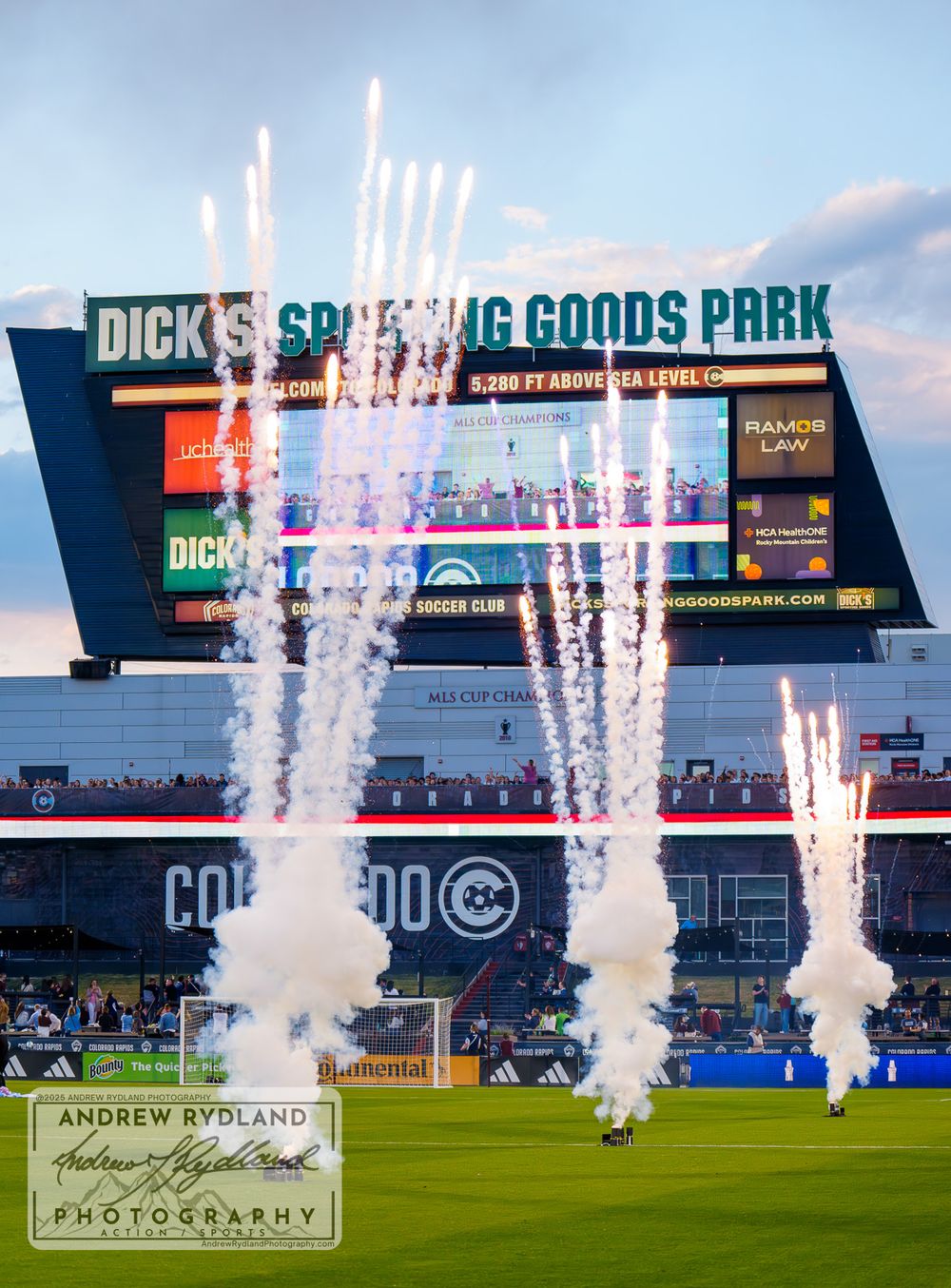 Fireworks display shooting up in front of Dick's Sporting Goods Park stadium scoreboard at dusk.