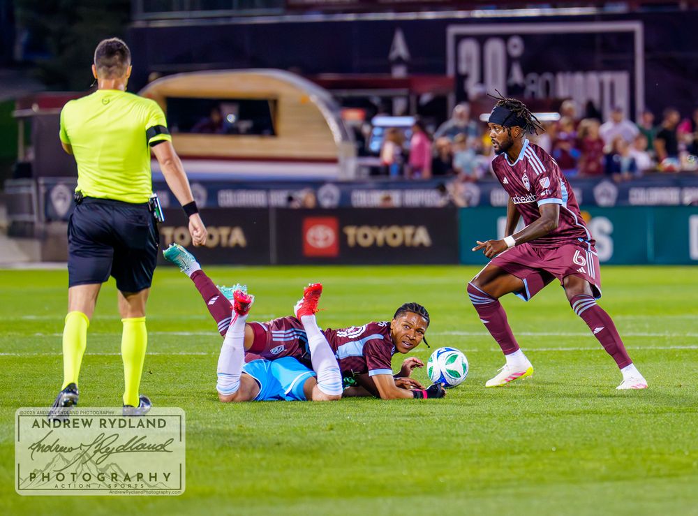 Soccer players scramble for loose ball with referee watching nearby.