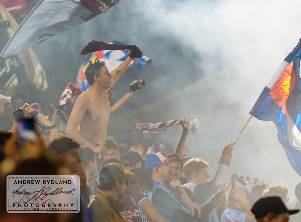 Soccer fans celebrate in a smoke-filled stadium with flags and raised arms during an intense match moment.