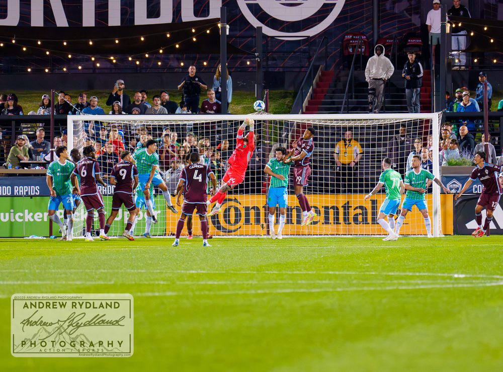 Soccer players jump to defend a goal during a nighttime match under stadium lights.