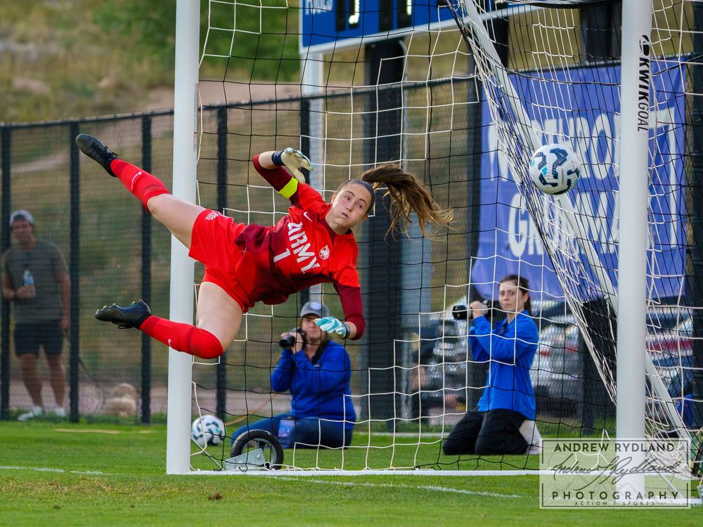 A goalkeeper in red makes an acrobatic diving save at the goalpost during a match.