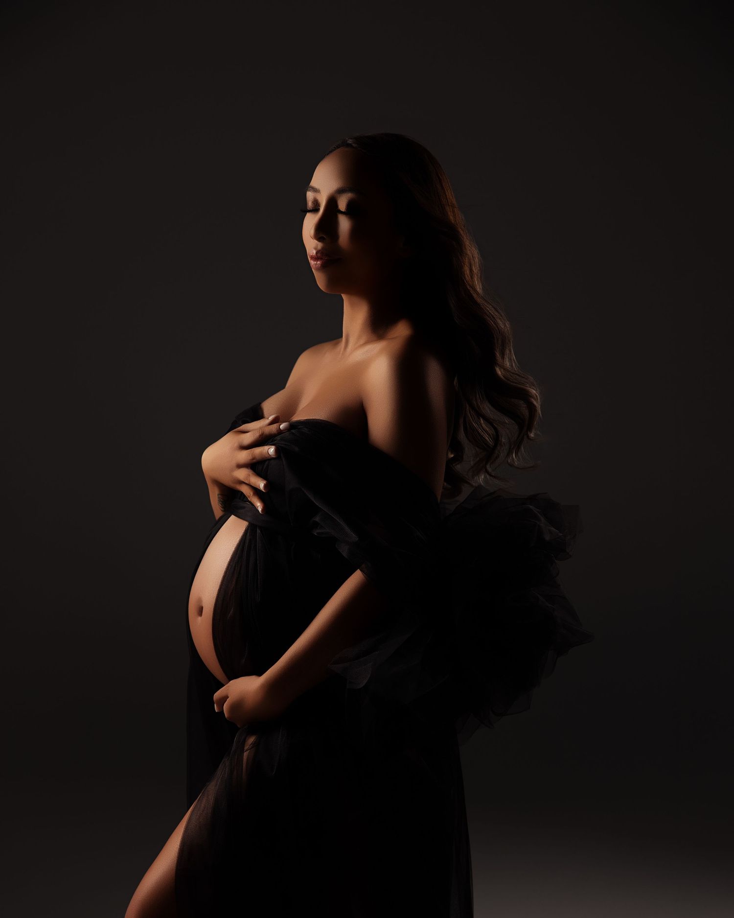 Artistic maternity portrait with dramatic lighting showing an expectant mother in a black tulle gown against dark backdrop.