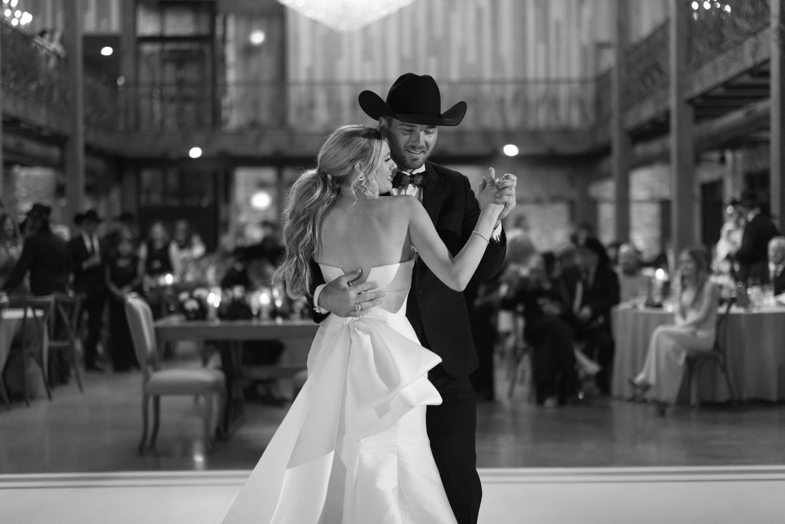 A couple dances elegantly at a wedding reception, with the bride in a flowing white dress and partner in black cowboy hat.