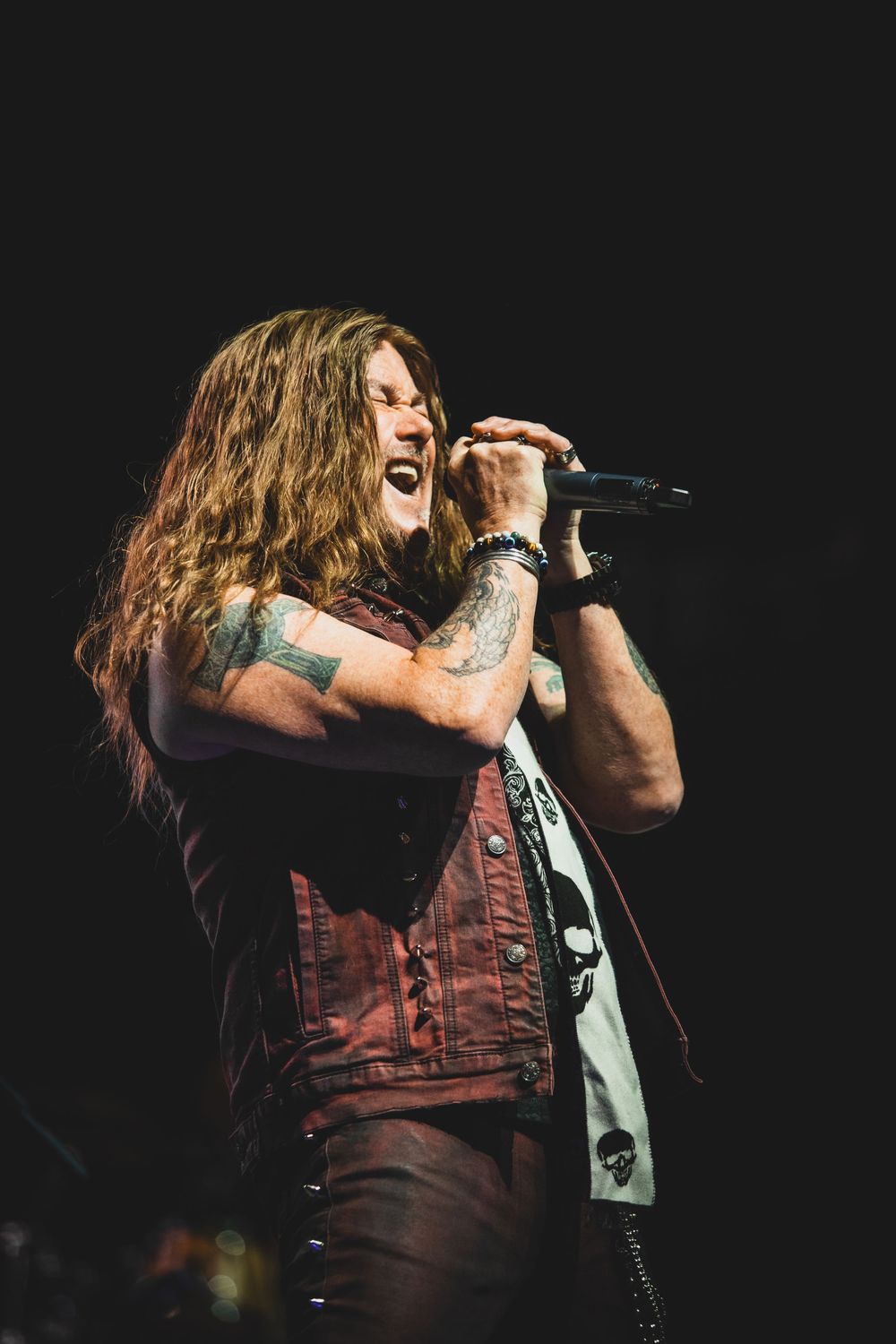 Rock singer with long wavy hair and tattoos performs passionately on stage in brown leather vest during concert.