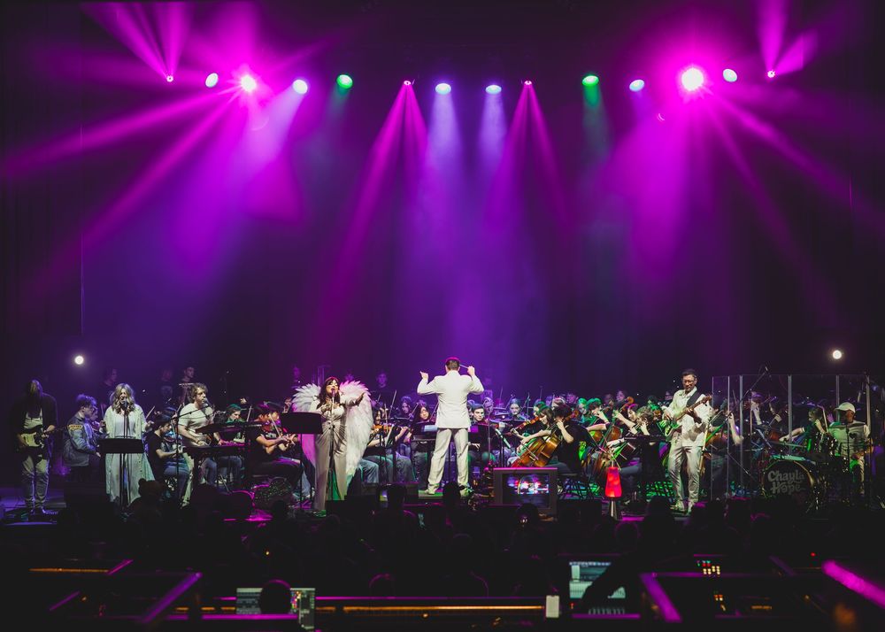 Concert stage bathed in vibrant purple and pink lighting with performers and orchestra silhouettes creating dramatic ambiance.