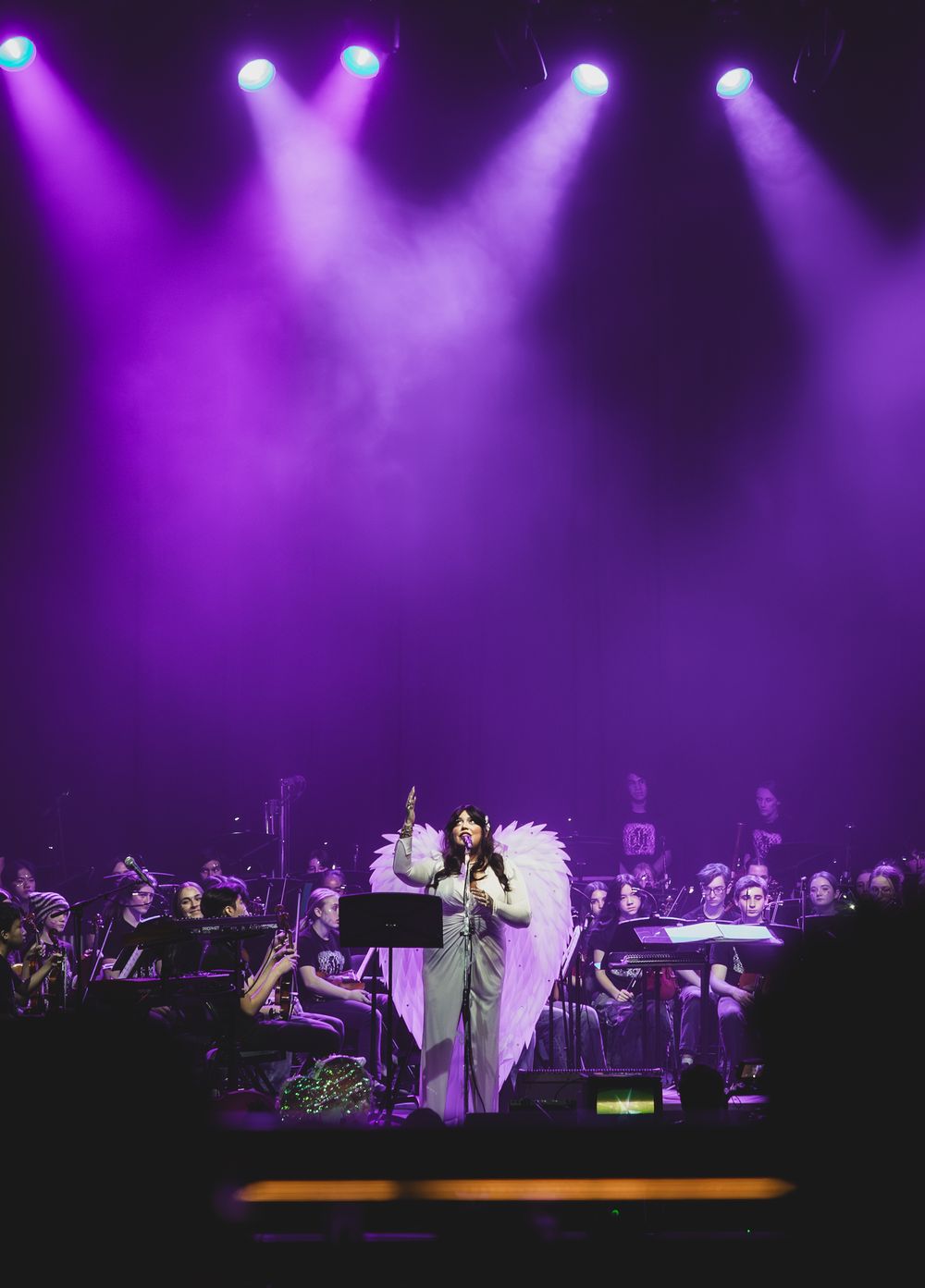 Wide stage performance scene illuminated by dramatic purple spotlights with orchestra and performers in silhouette.