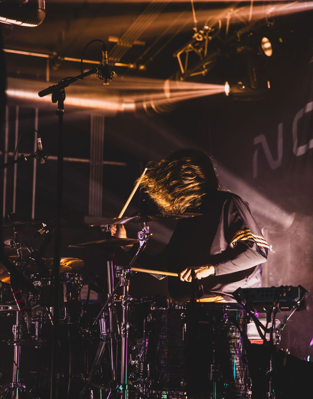 A drummer performs on stage during a concert with dramatic lighting and shadows creating a dynamic atmosphere.