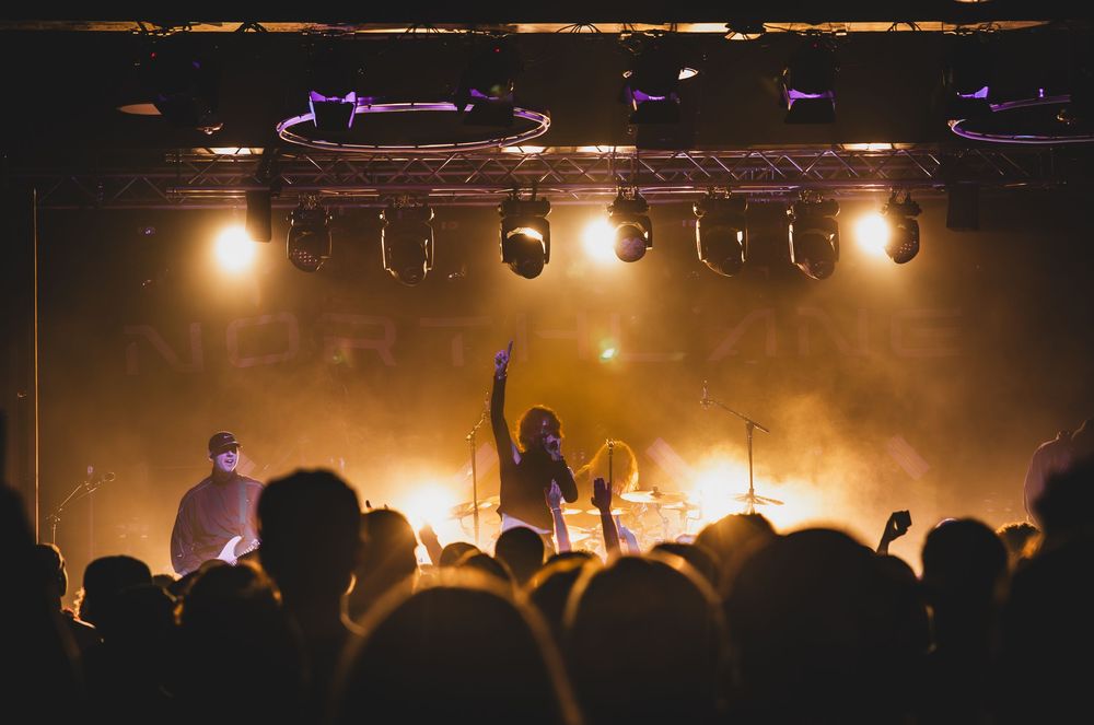 Concert crowd in silhouette stands with arms raised under stage lights during a live performance at a music venue.