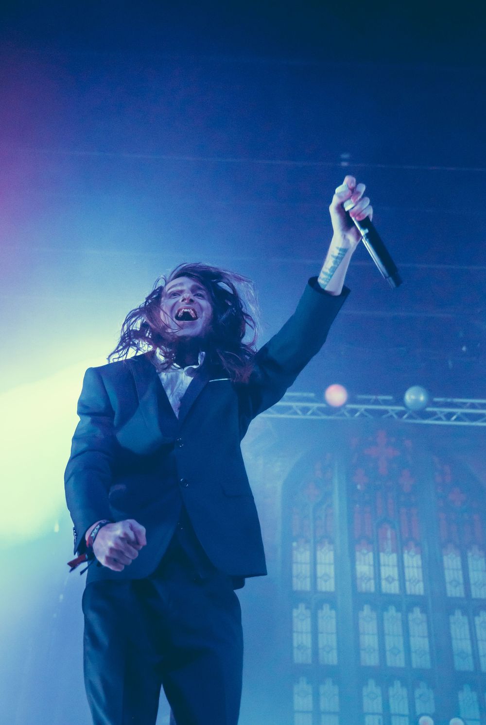 A performer in a dark suit expressively moves and dances against a moody blue-lit stage background at a concert.