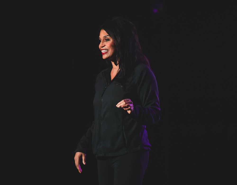 Performer dressed in black clothing stands on darkened stage with purple lighting effect in background.