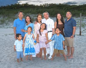A group stands together on a sandy beach during sunset wearing coordinated blue and white outfits for a family photo.